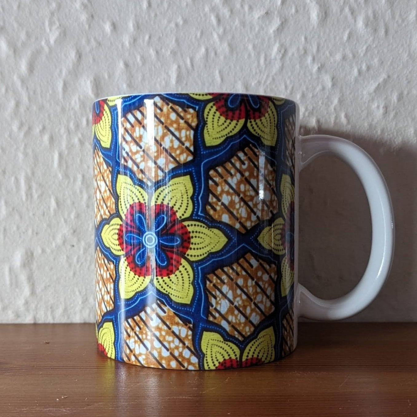 A white ceramic mug with a bold geometric flower pattern. The flower is an African Daisy and the colours are yellow red & blue. It has a shiny gloss glaze.