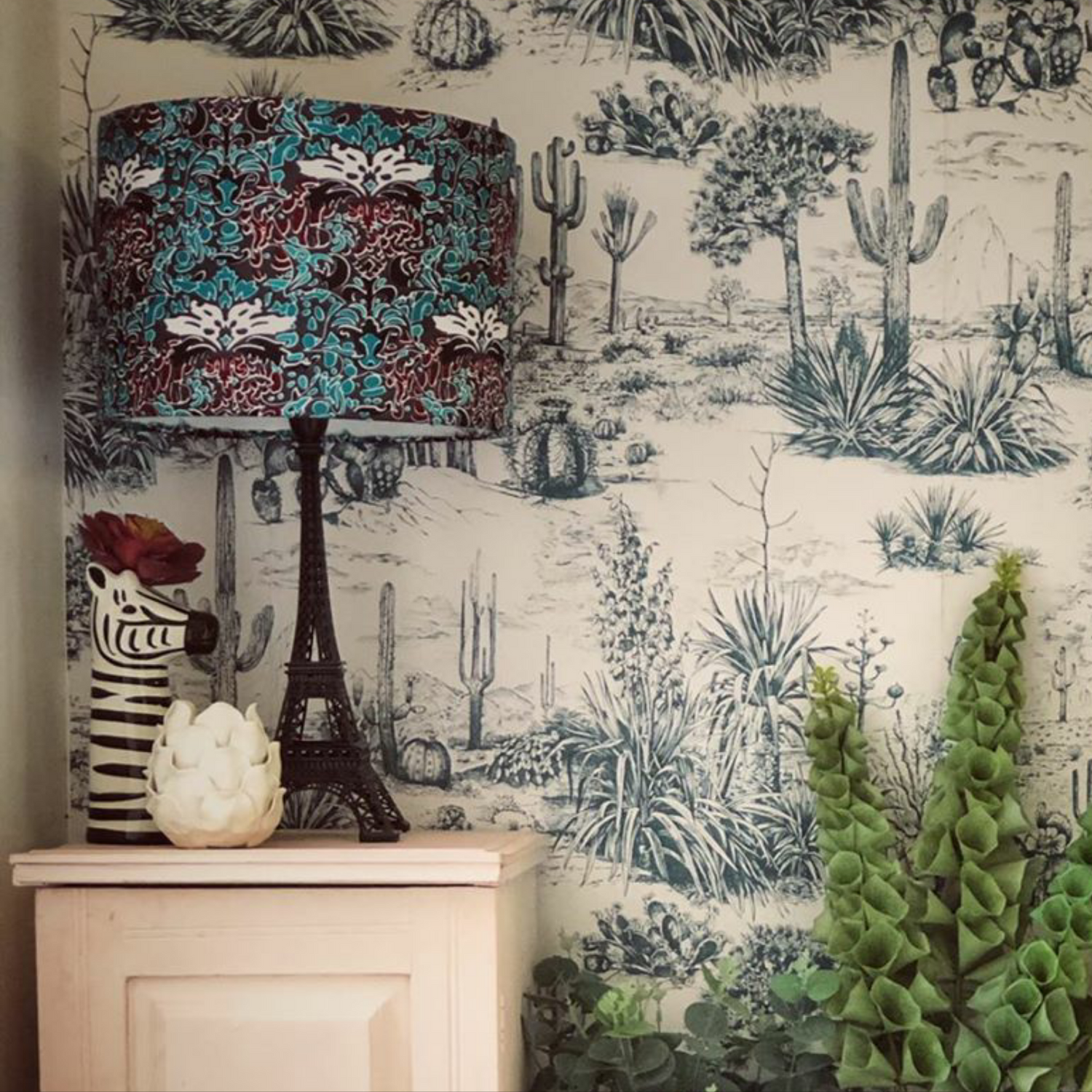 Mint red wine wild west style African drum lampshade sitting on a corner table with other decorative ornaments. It is against a background with a cactus themed botanical wallpaper feature wall. The lampshade is paired with a complimenting sleek black lamp base. 