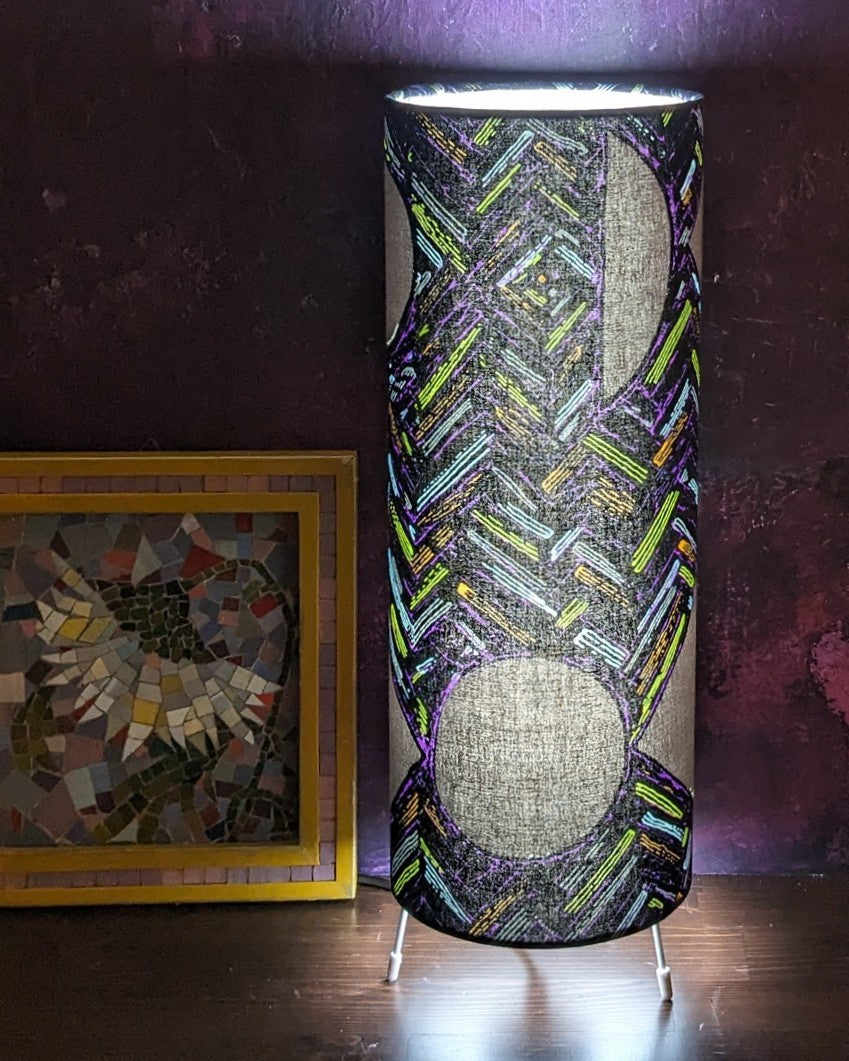 Tall slim celestial night sky inspired free standing table lamp. Made in a patterned African wax print in pastel blue green purple grey on a blackish-grey colourway.. Lamp is switched on and the light brightens up the pattern beautifully. 