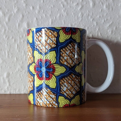 A white ceramic mug with a bold geometric flower pattern. The flower is an African Daisy and the colours are yellow red & blue. It has a shiny gloss glaze.