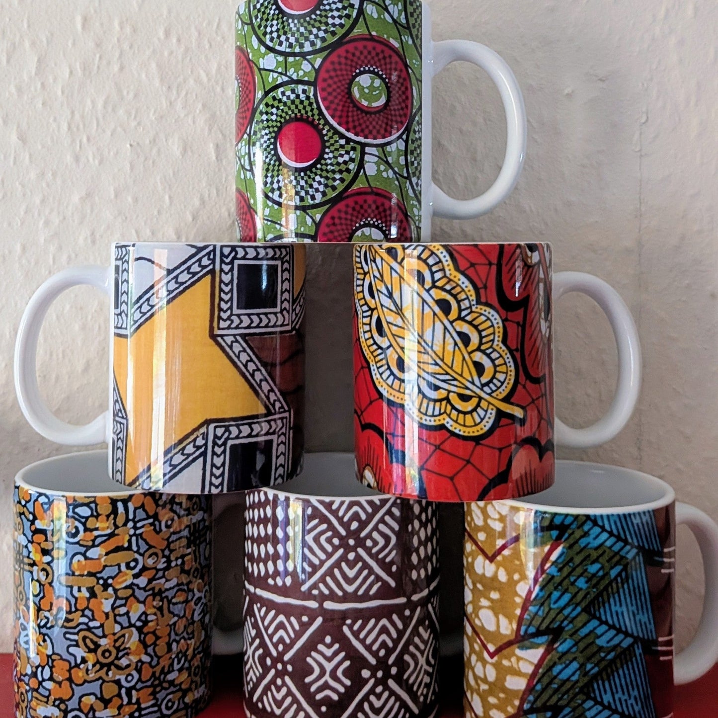 Stack of colorful African wax print patterned ceramic mugs on a red surface with a white wall background