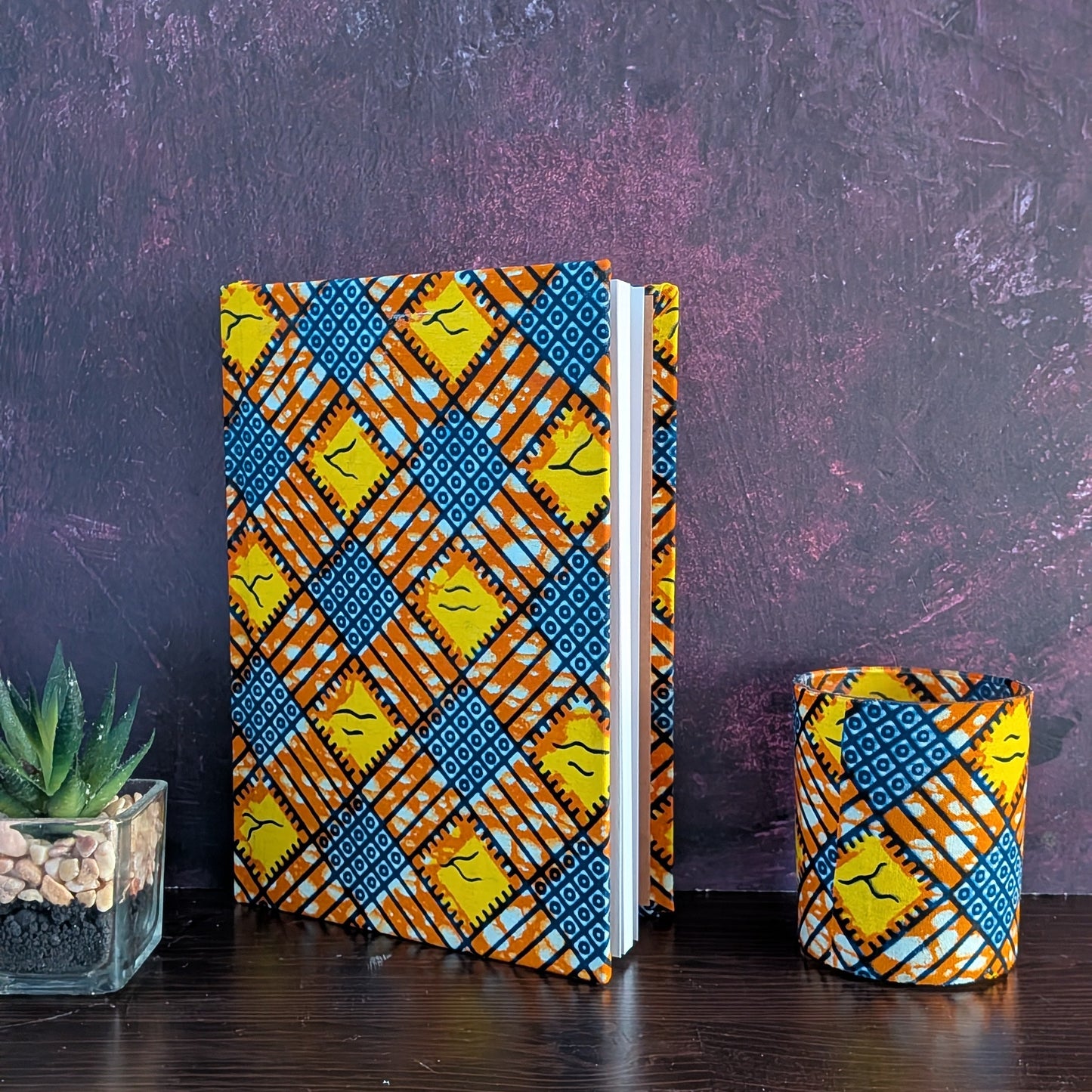 Notebook and candle holder with colorful criss cross geometric block pattern on a dark surface and purple background