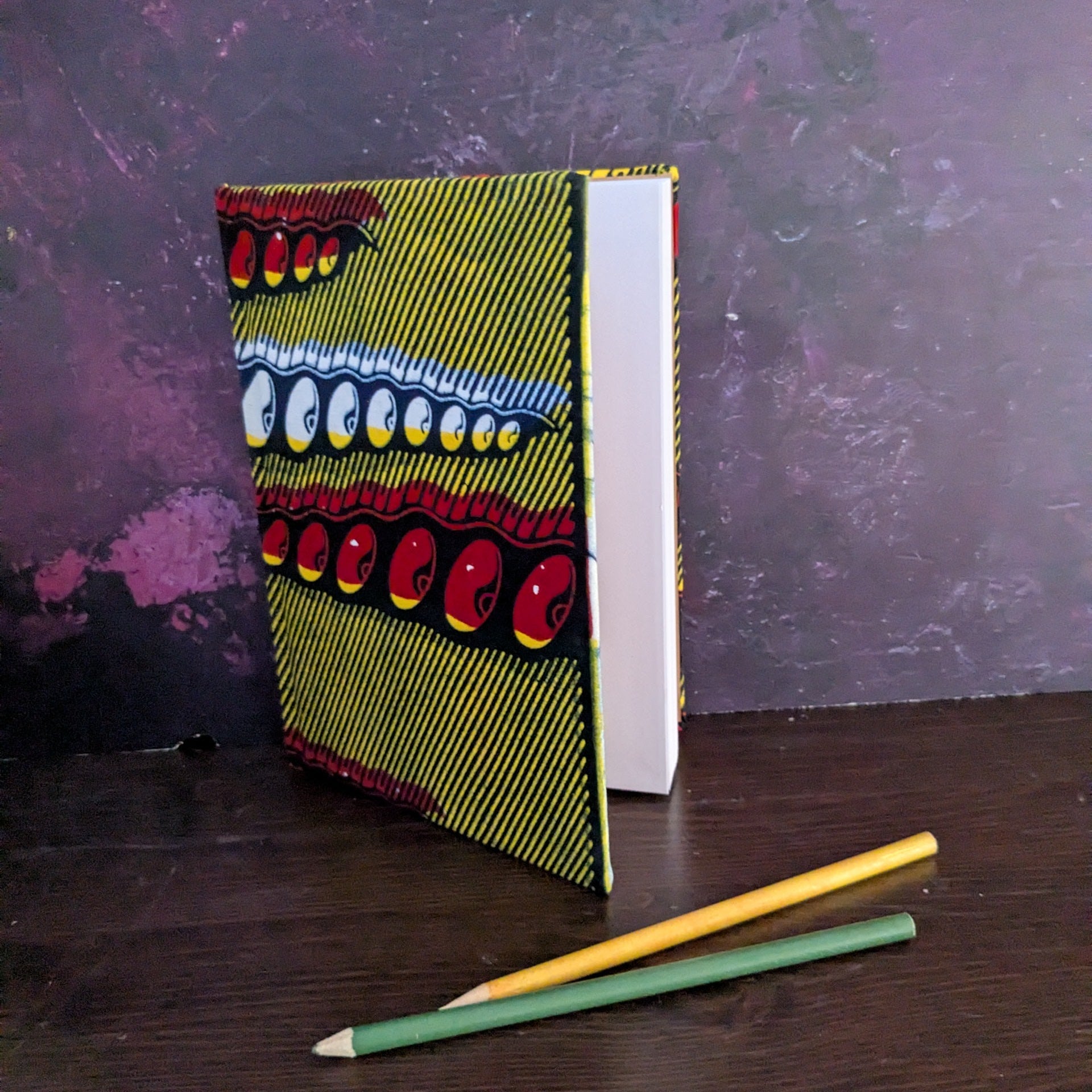 Notebook with a vibrant African wax print design on a dark surface with two pencils. 