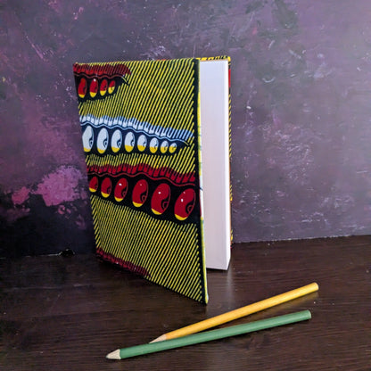 Notebook with a vibrant African wax print design on a dark surface with two pencils. 