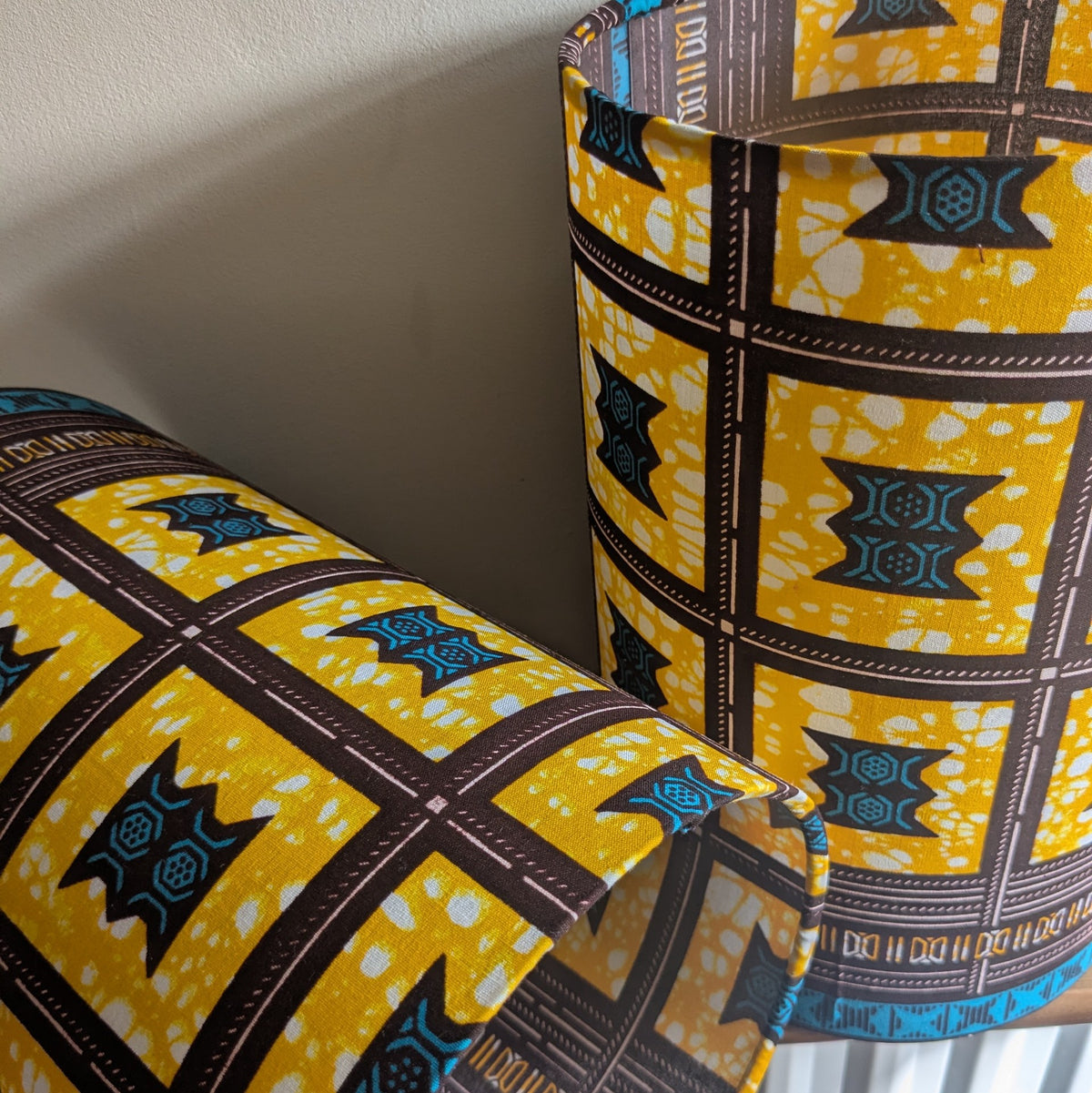 A close-up of a set of cylindrical lampshades made with in bold African wax print fabric. They have a yellow, brown and teal patterned design. A second matching wrapped item is partially visible in the foreground against a plain light wall.
