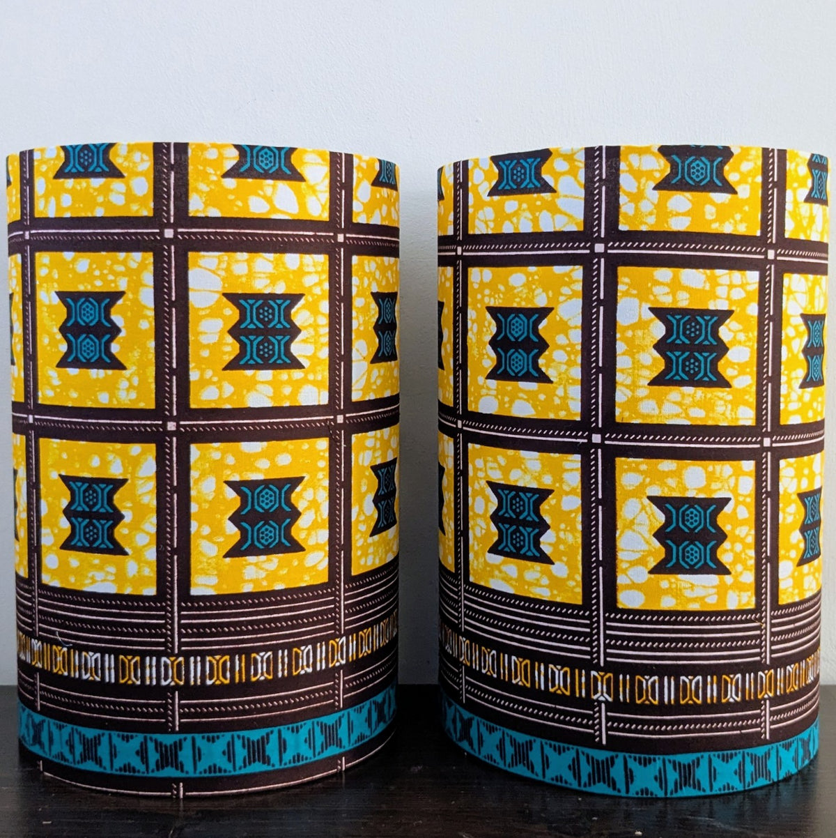 Two cylindrical lampshades featuring a bold yellow, brown, and teal African wax print geometrical pattern. They are standing side-by-side on a dark wooden surface against a plain white wall.