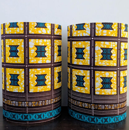 Two cylindrical lampshades featuring a bold yellow, brown, and teal African wax print geometrical pattern. They are standing side-by-side on a dark wooden surface against a plain white wall.
